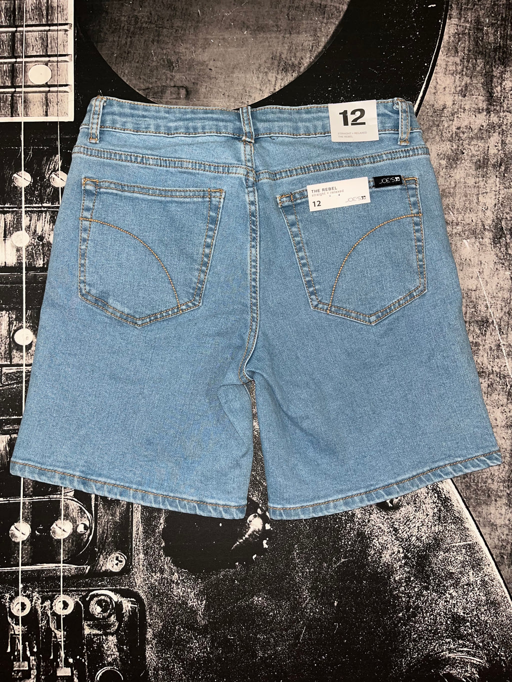 Light blue denim shorts shown from the back, laid flat on a black-and-white guitar-themed background. The shorts have a 12-inch waist, two back pockets with decorative stitching, and visible size labels reading 12.