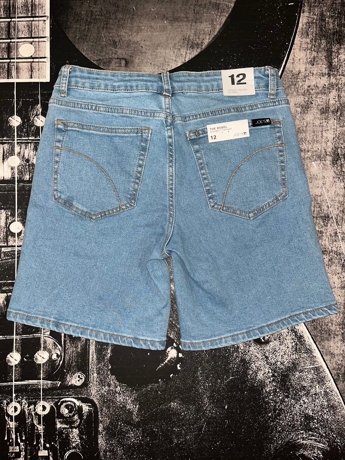 Light blue denim shorts shown from the back, laid flat on a black-and-white guitar-themed background. The shorts have a 12-inch waist, two back pockets with decorative stitching, and visible size labels reading 12.