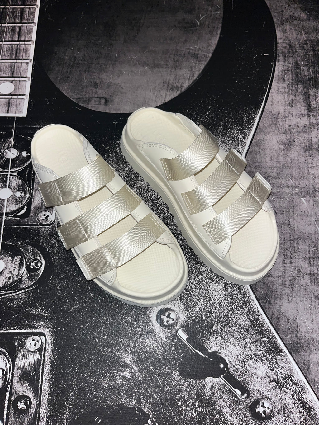 A pair of platform slide sandals with wide, woven-look straps in a light metallic champagne color, displayed on a textured black and gray guitar body.