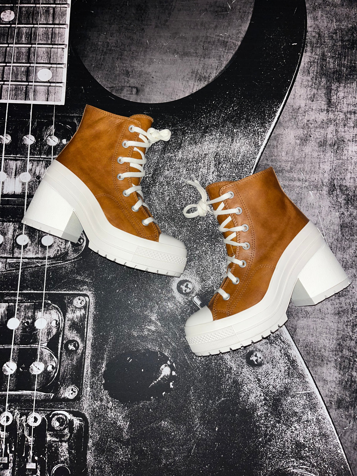A pair of brown platform ankle boots with chunky white block heels and white laces, styled on a black electric guitar against a textured gray background.