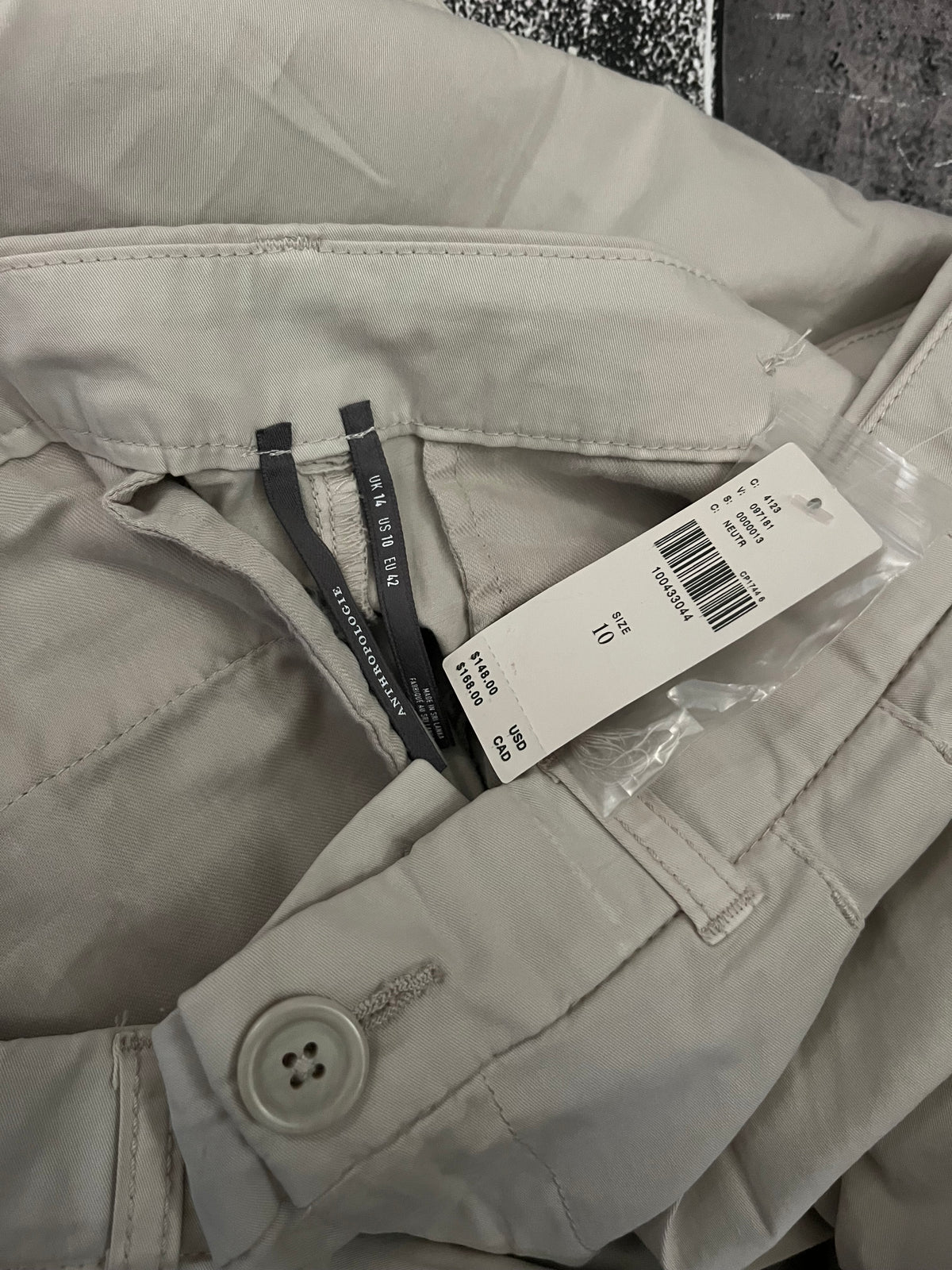 A close-up of a light beige or off-white garment, likely a jacket or coat, showing the collar, buttoned cuff, and attached white size tag with a barcode.
