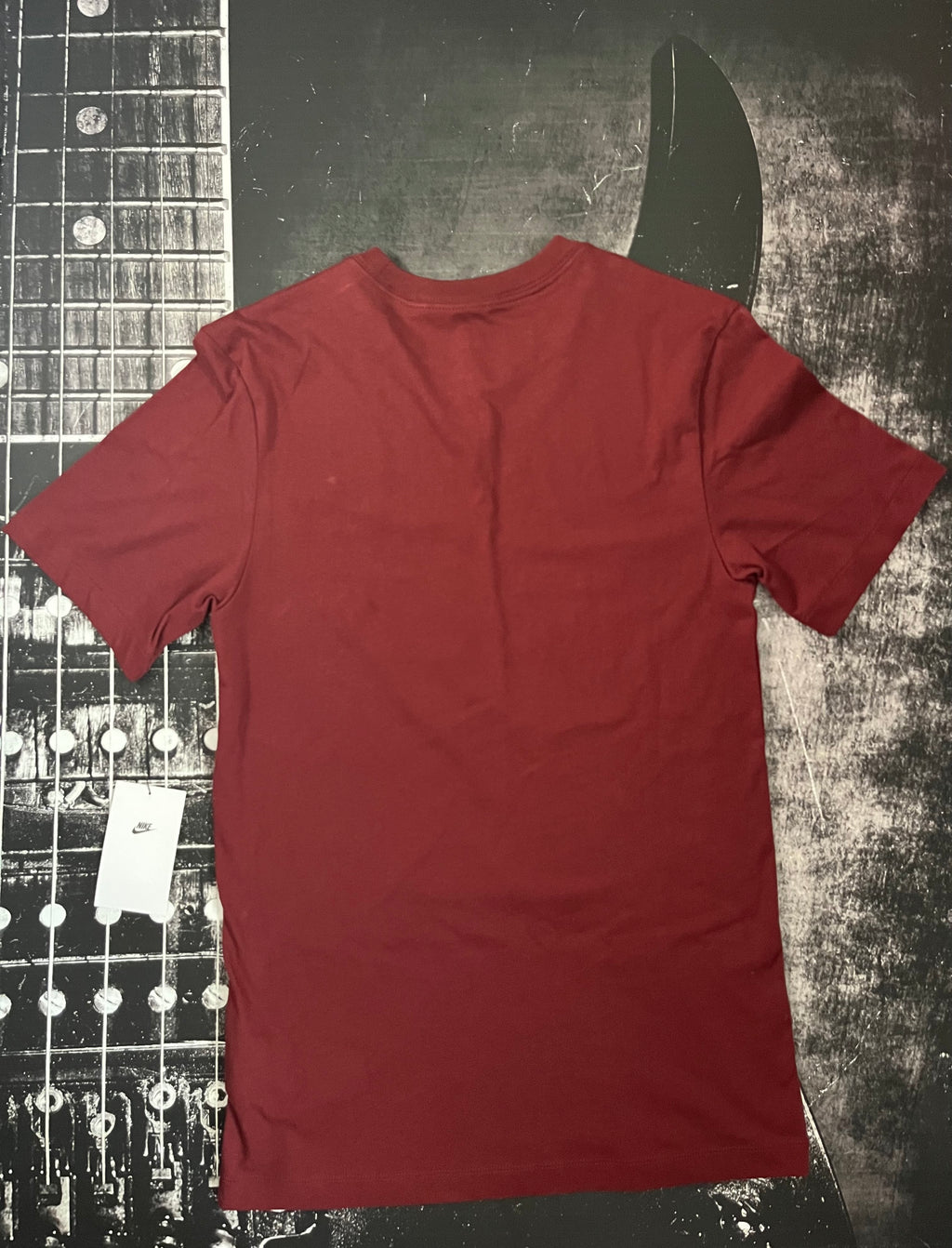 A plain short-sleeve crewneck t-shirt in a deep burgundy red, laid flat on a dark textured background with a guitar headstock graphic.