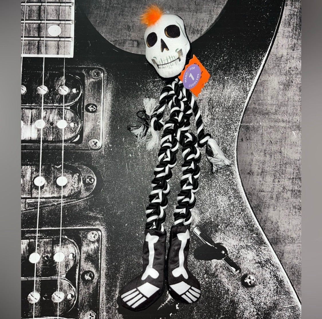 A Halloween skeleton doll with a white skull head, black and white striped legs, and orange hair, posed on top of a black electric guitar.