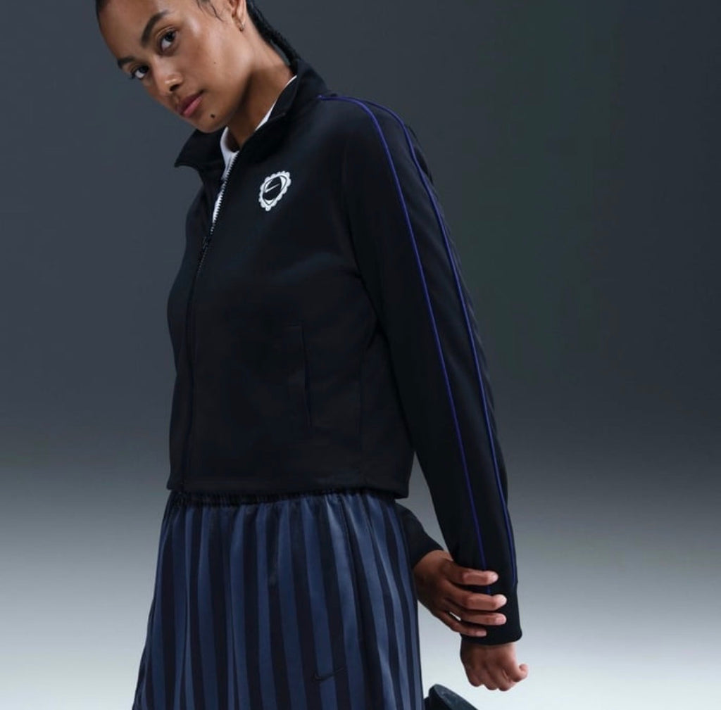 A woman wearing a black athletic zip-up jacket with blue stripe details and a matching pleated skirt, photographed against a dark gray studio background.
