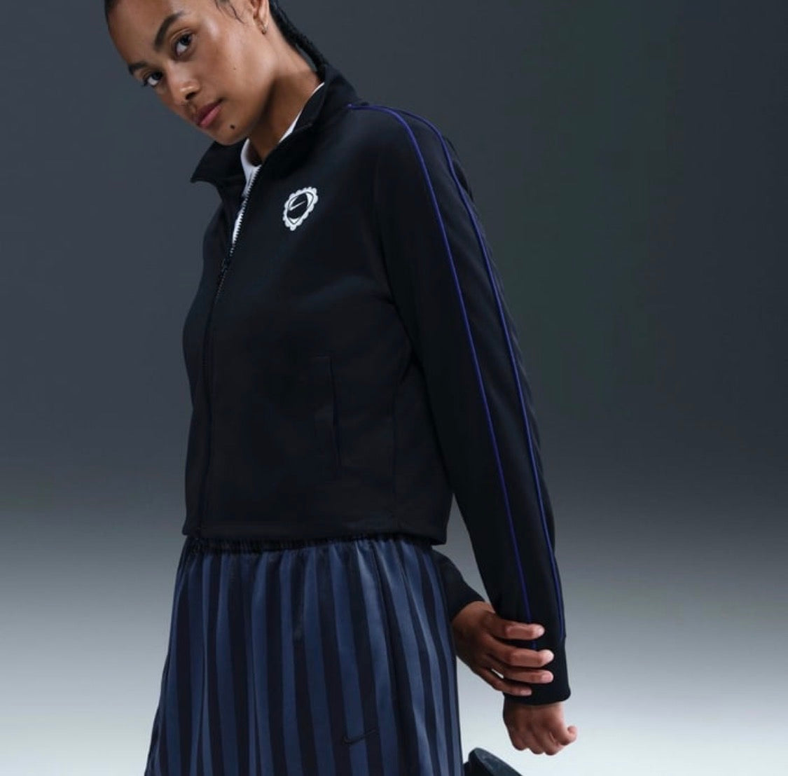 A woman wearing a black athletic zip-up jacket with blue stripe details and a matching pleated skirt, photographed against a dark gray studio background.