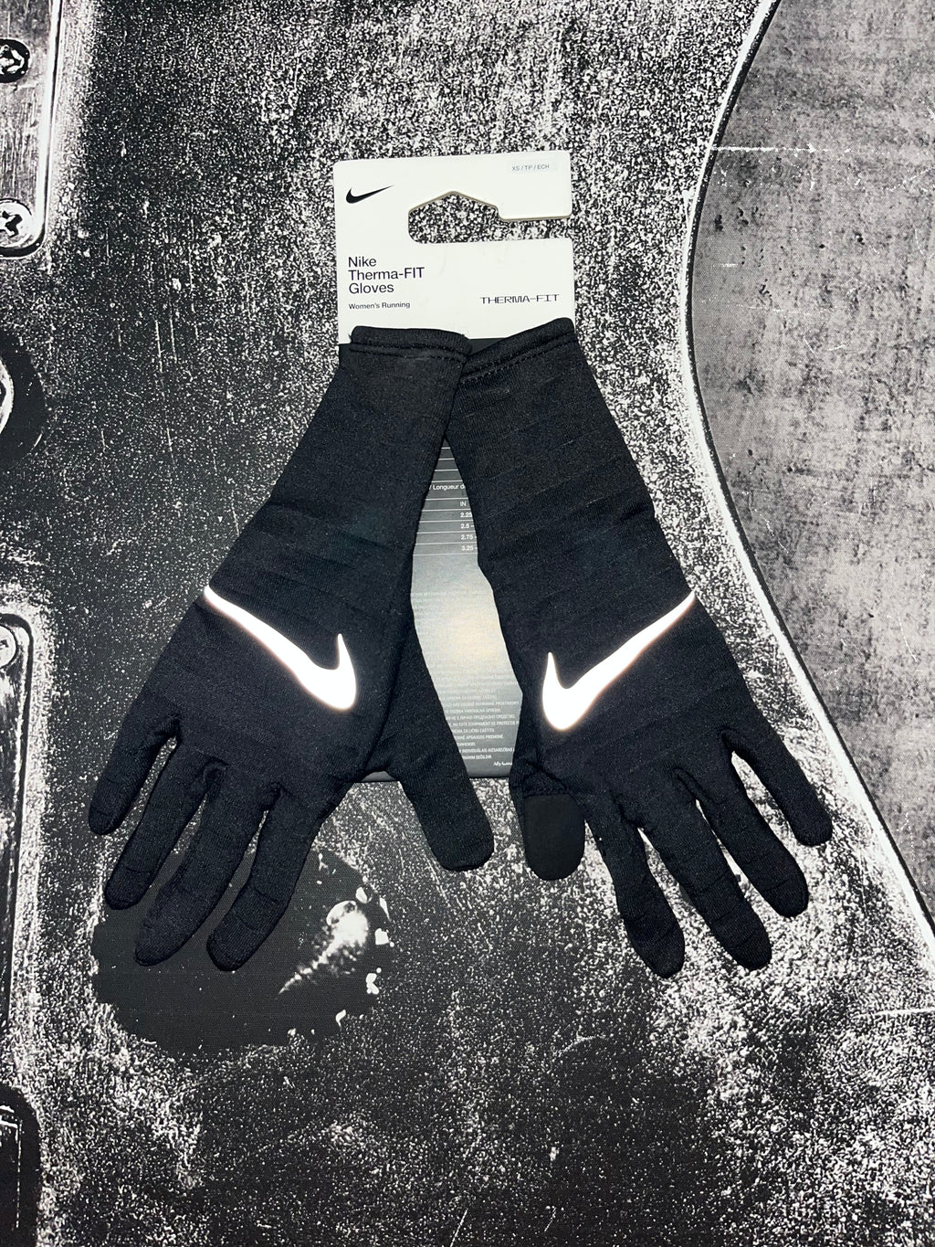 A pair of black Nike full-finger gloves with reflective white swooshes, shown with their original white retail hang tag on a textured, dark metallic surface.