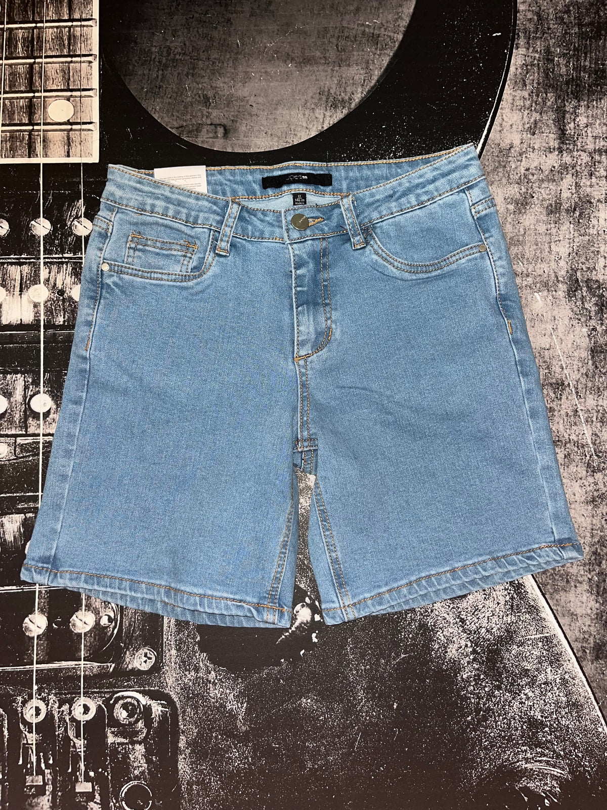 Light blue denim shorts with a front split seam, laid flat on a black and white guitar-patterned background.