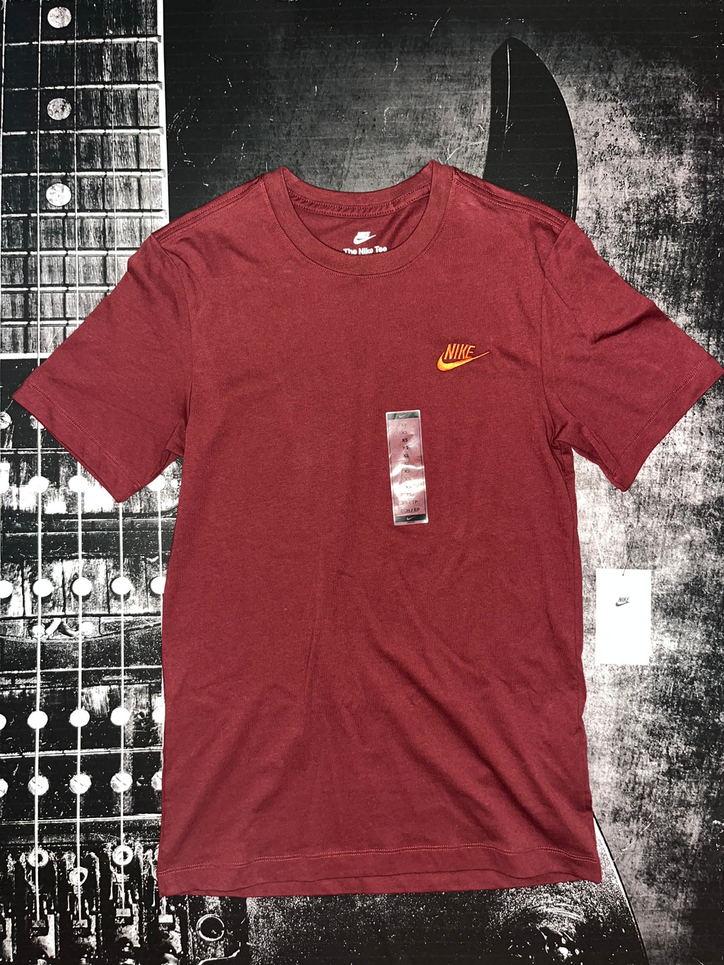A maroon short-sleeve Nike t-shirt with a small yellow Swoosh logo on the left chest, displayed flat against a black and white guitar headstock background.