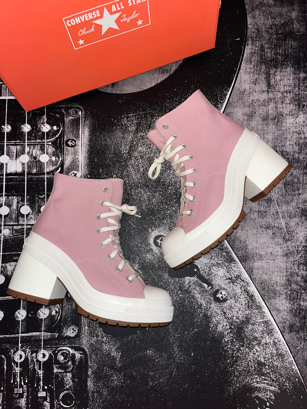 A pair of Converse All Star platform ankle boots in dusty pink canvas with white laces, white rubber toe caps and midsoles, and brown gum outsoles. An orange Converse shoebox is partially visible at the top, and the boots are styled on a black electric guitar against a dark textured background.