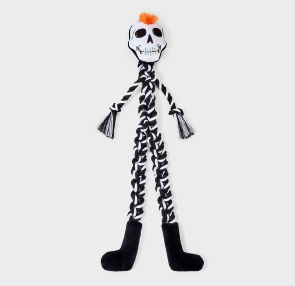 A skeleton-themed dog toy with a white skull head, black braided body, black feet, and a small orange tuft on top, shown against a light gray background.