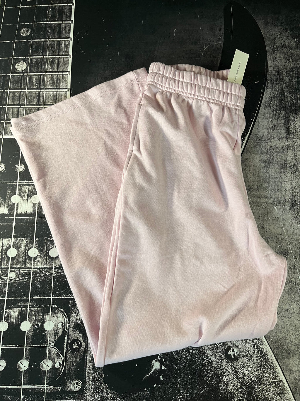 Light pink sweatpants folded on a dark textured surface with a guitar headstock in the background.