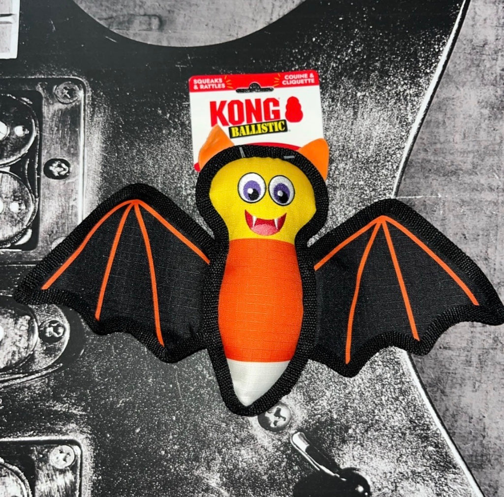 A Kong Ballistic bat-shaped dog toy with black wings, orange and white candy corn body, and a yellow face, shown with its retail packaging against a black guitar background.