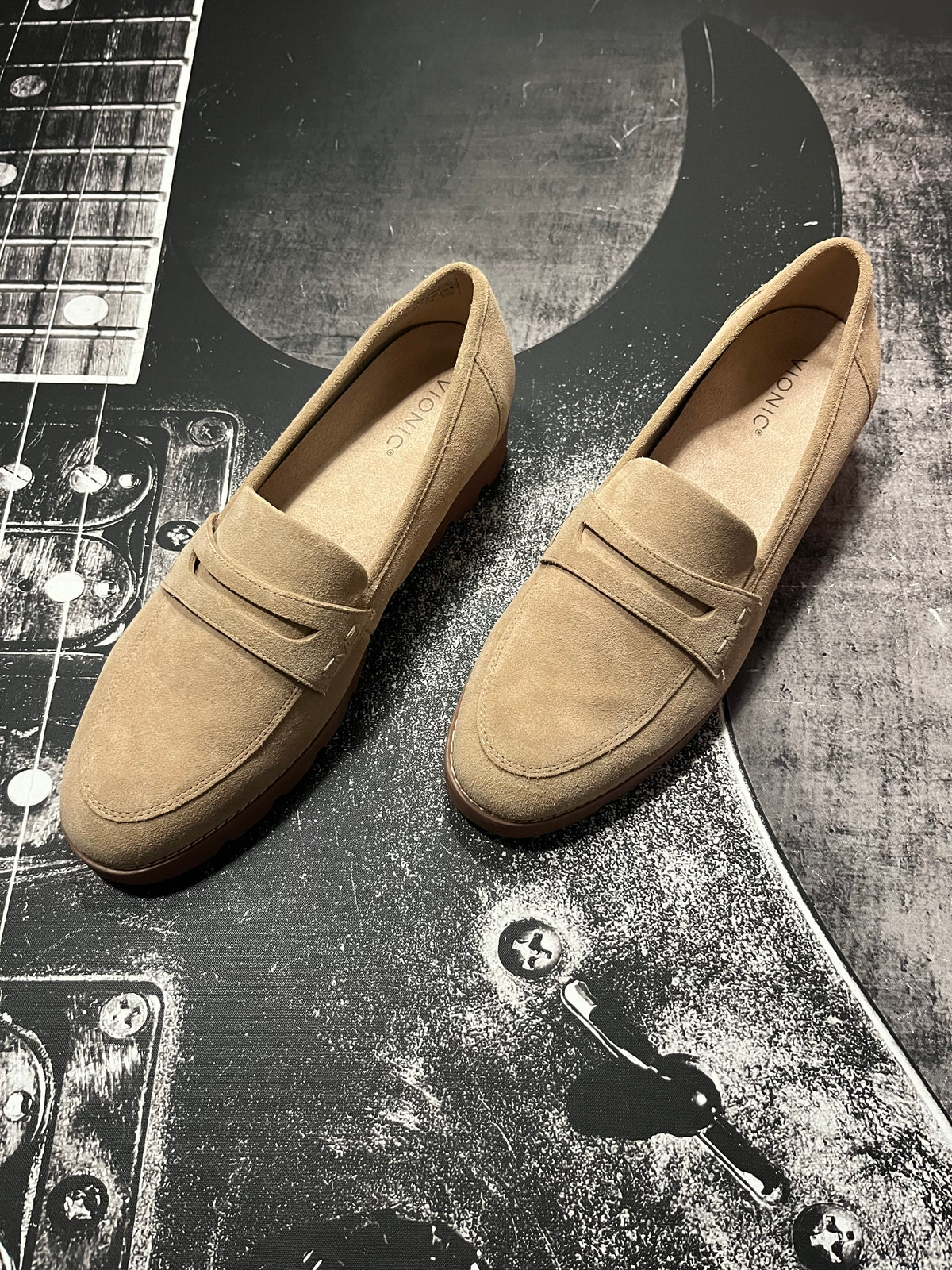 A pair of tan suede penny loafers with low block heels, displayed on a black and white guitar-themed background.