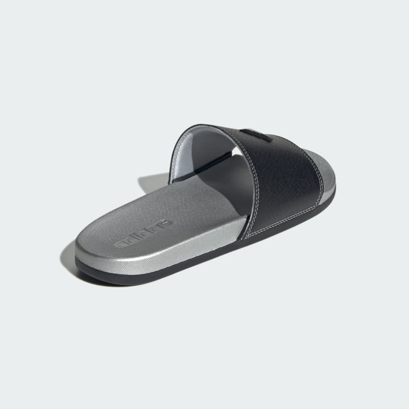 A single slide sandal shown from a three-quarters perspective on a light gray background. The sandal features a textured black upper strap and a metallic silver-gray footbed with the embossed brand name 'MILA'.