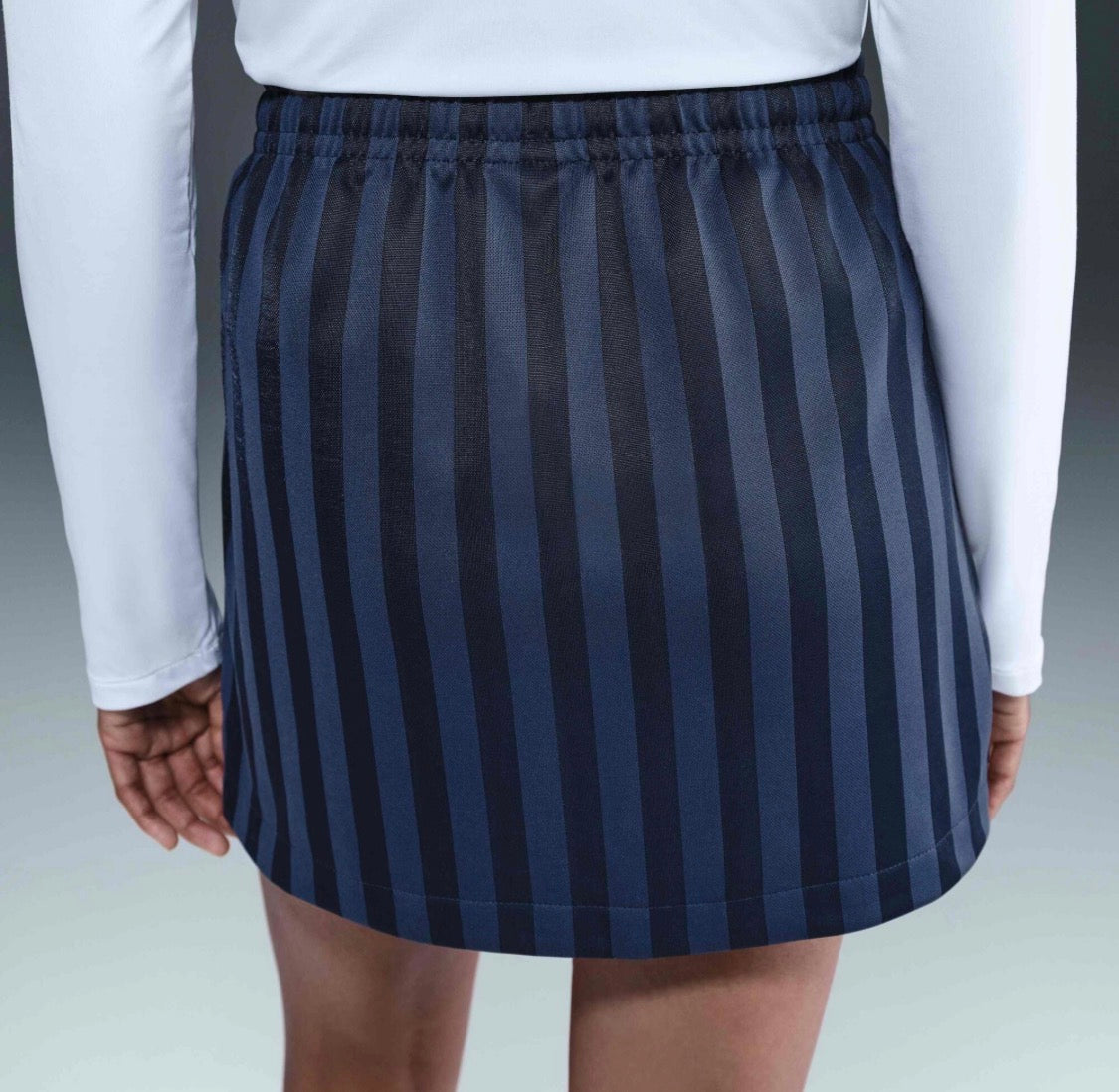 A close-up back view of a person wearing a short athletic skirt with vertical navy and dark gray stripes, paired with a white long-sleeve top.