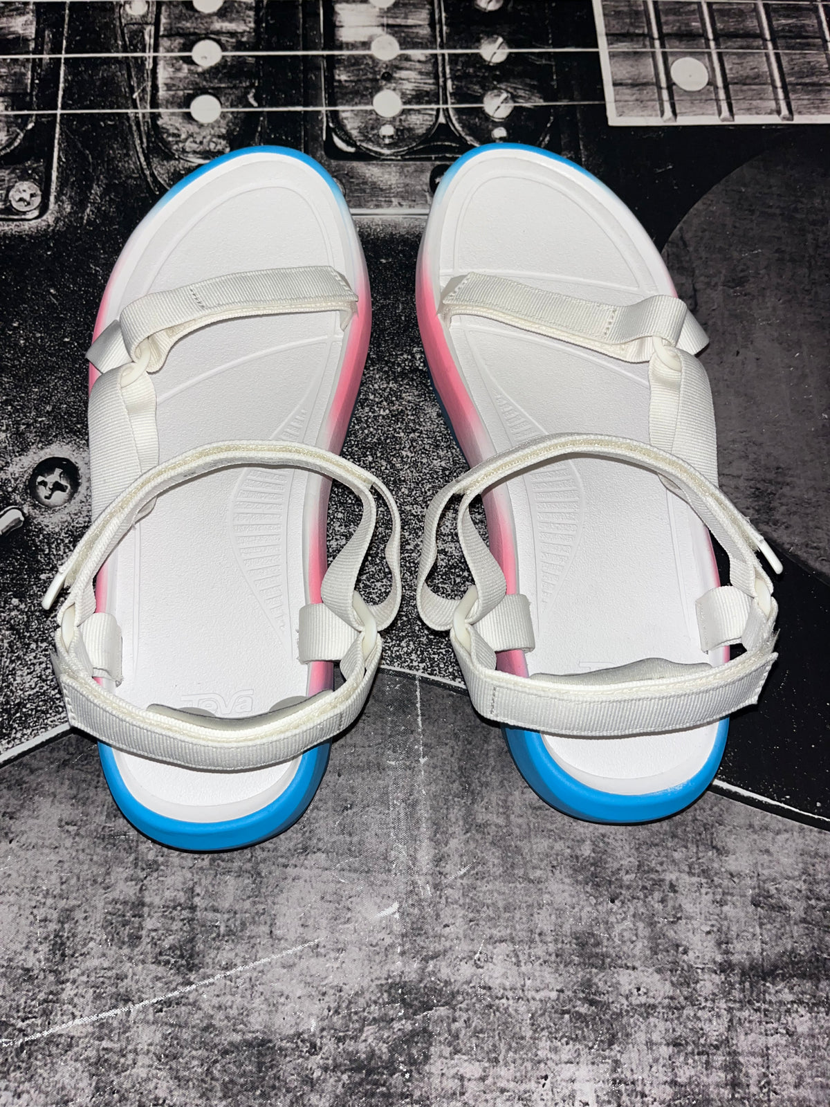 A pair of white platform sandals with white straps, pink sides, and blue soles, displayed on a dark textured surface with a guitar headstock in the background.