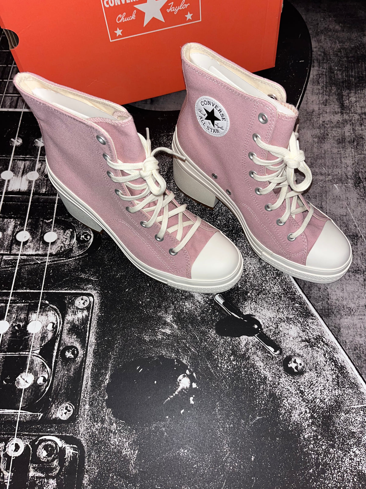 A pair of Converse high-top boots in dusty pink canvas with white laces, white rubber toe caps, and white mid-height heels, displayed next to their orange Converse box on a black guitar-print surface.