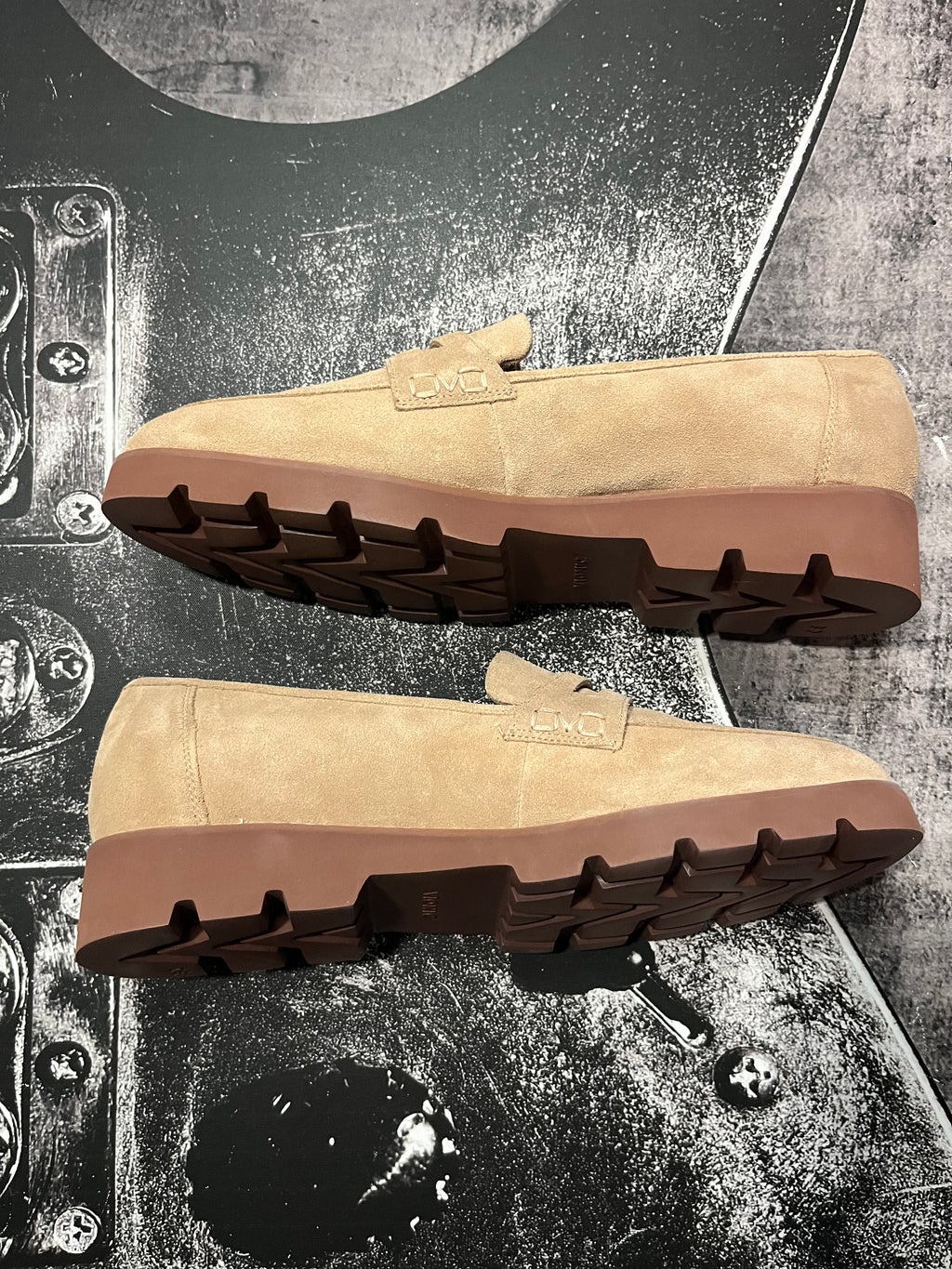 A pair of tan suede slip-on loafers with brown soles, displayed from the side and bottom angles on a textured black and gray background.