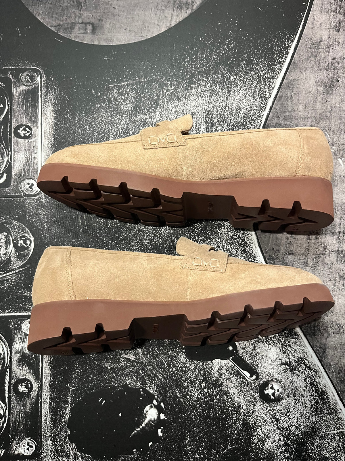 A pair of tan suede slip-on loafers with brown soles, displayed from the side and bottom angles on a textured black and gray background.