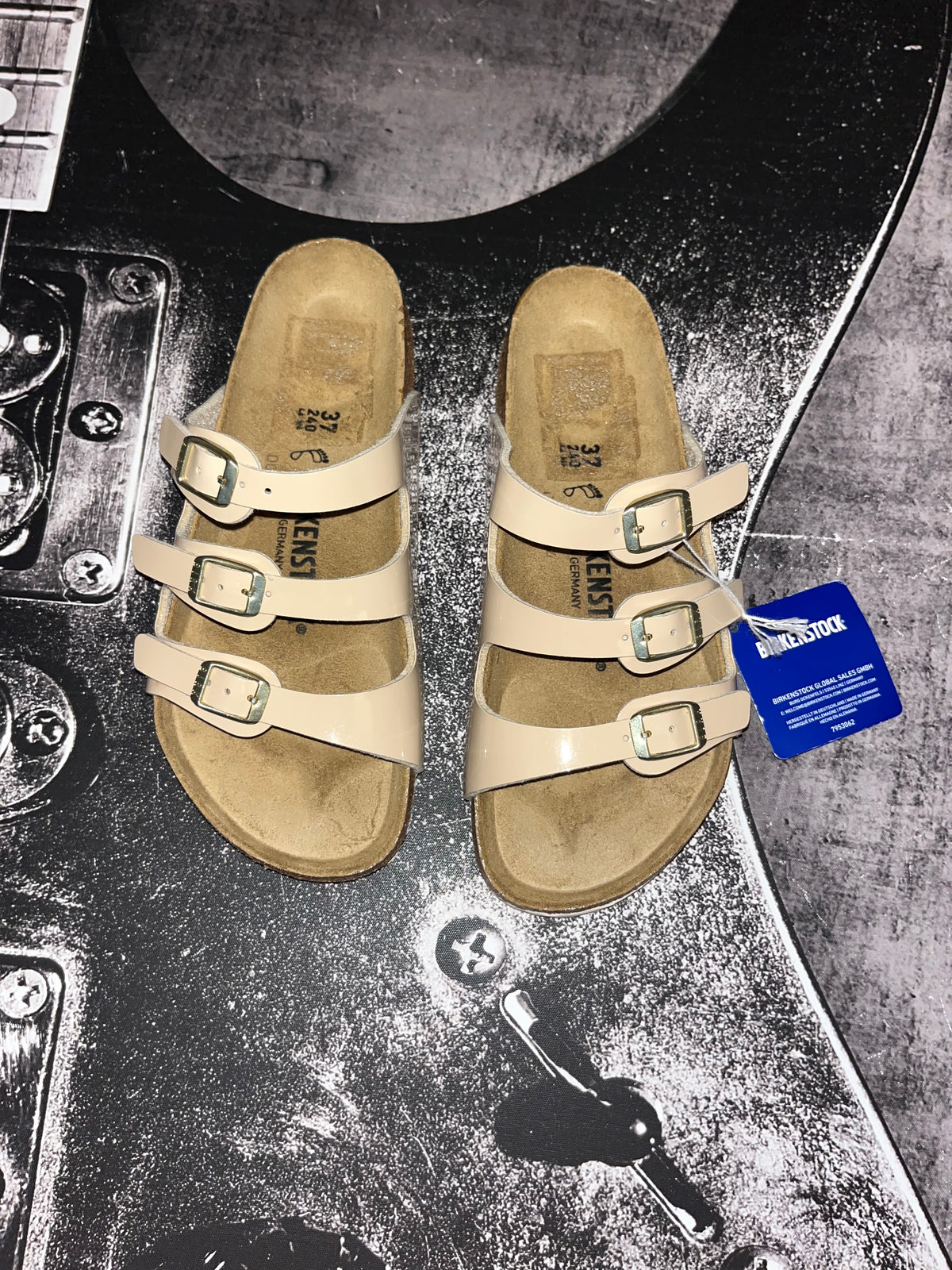 A pair of Birkenstock Arizona slide sandals in beige faux leather with adjustable buckled straps, shown on a dark, textured metal surface with a blue product tag attached.