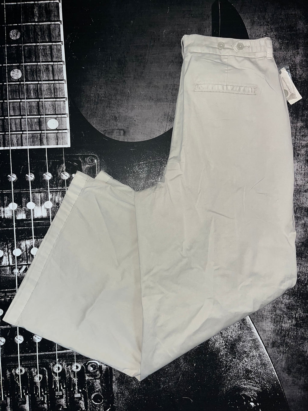A pair of light beige or off-white chino pants laid flat on a dark, textured background with a guitar headstock partially visible in the upper left corner.