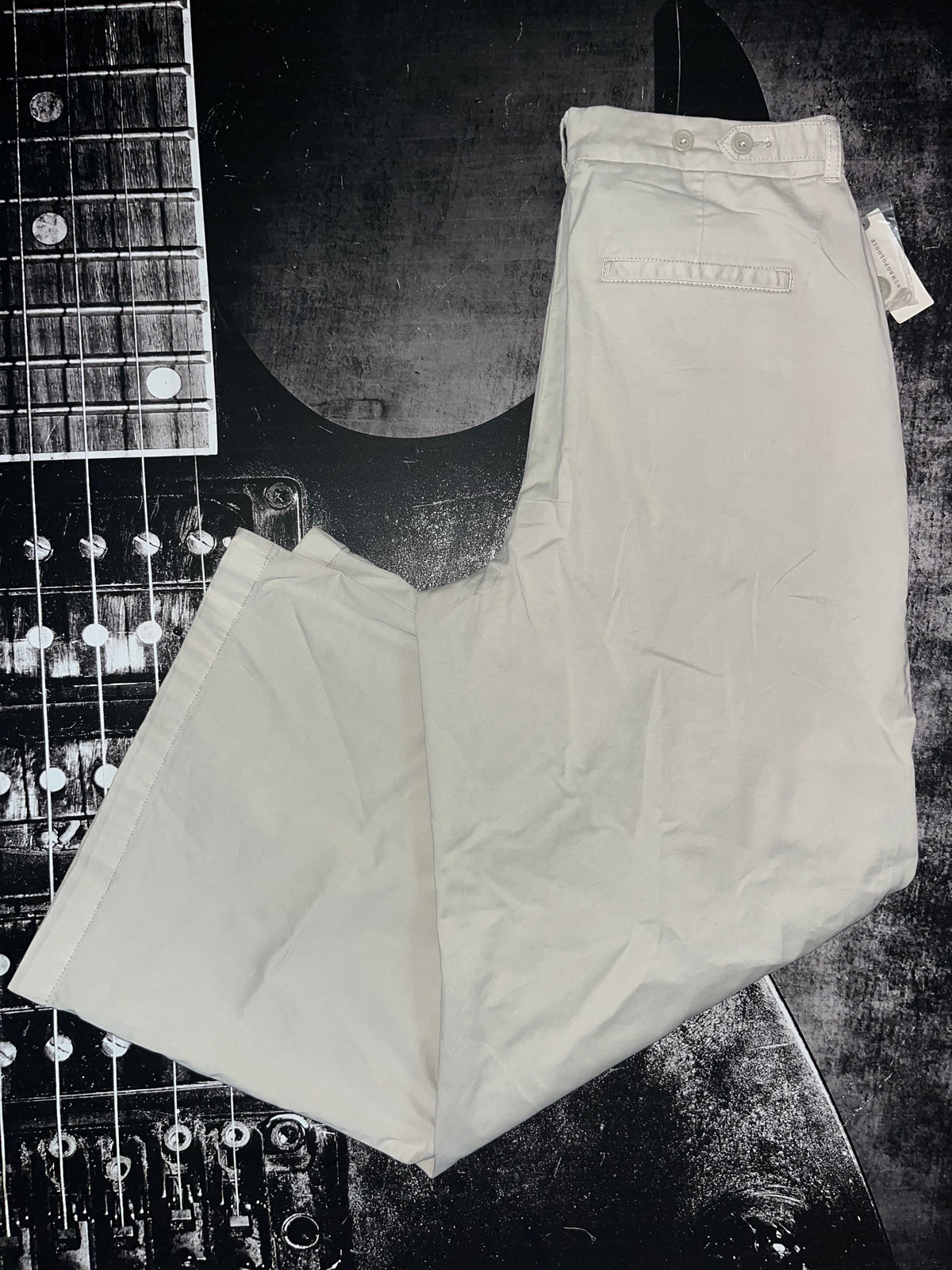 A pair of light beige or off-white chino pants laid flat on a dark, textured background with a guitar headstock partially visible in the upper left corner.