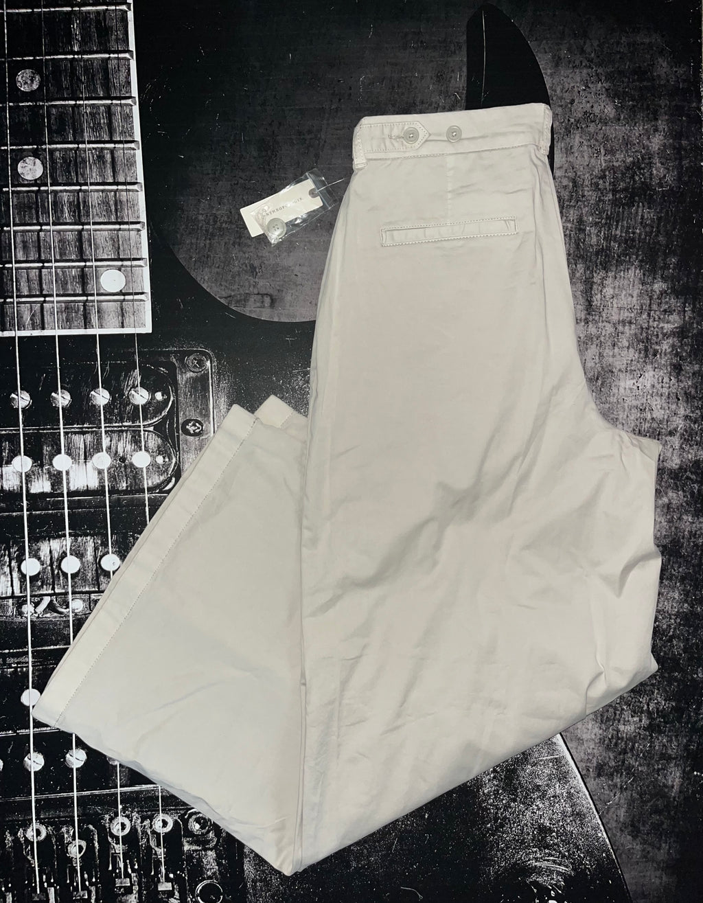 A pair of light beige or off-white trousers laid flat on a dark textured surface, partially overlaid by a guitar headstock and strings. No person, animal, or real environment is present, so this is Product/Packaging rather than Lifestyle.