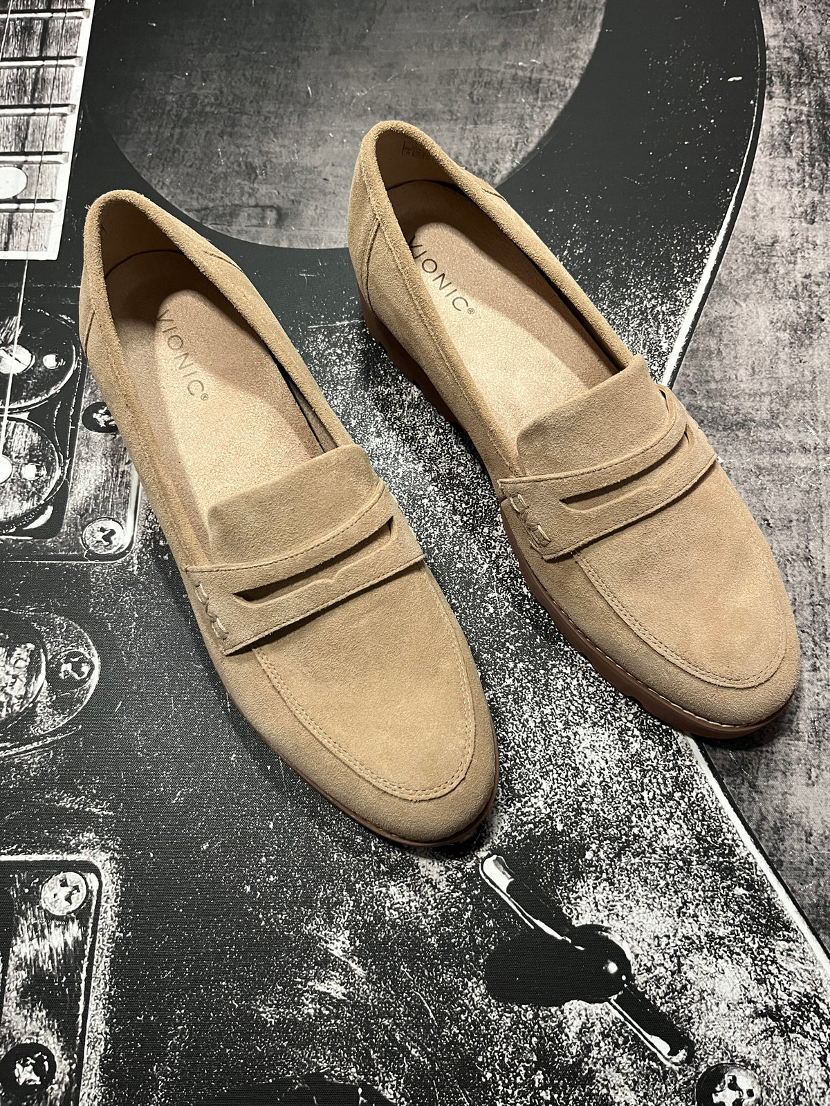 A pair of tan suede penny loafers with a low block heel, displayed on a dark, textured guitar headstock background.