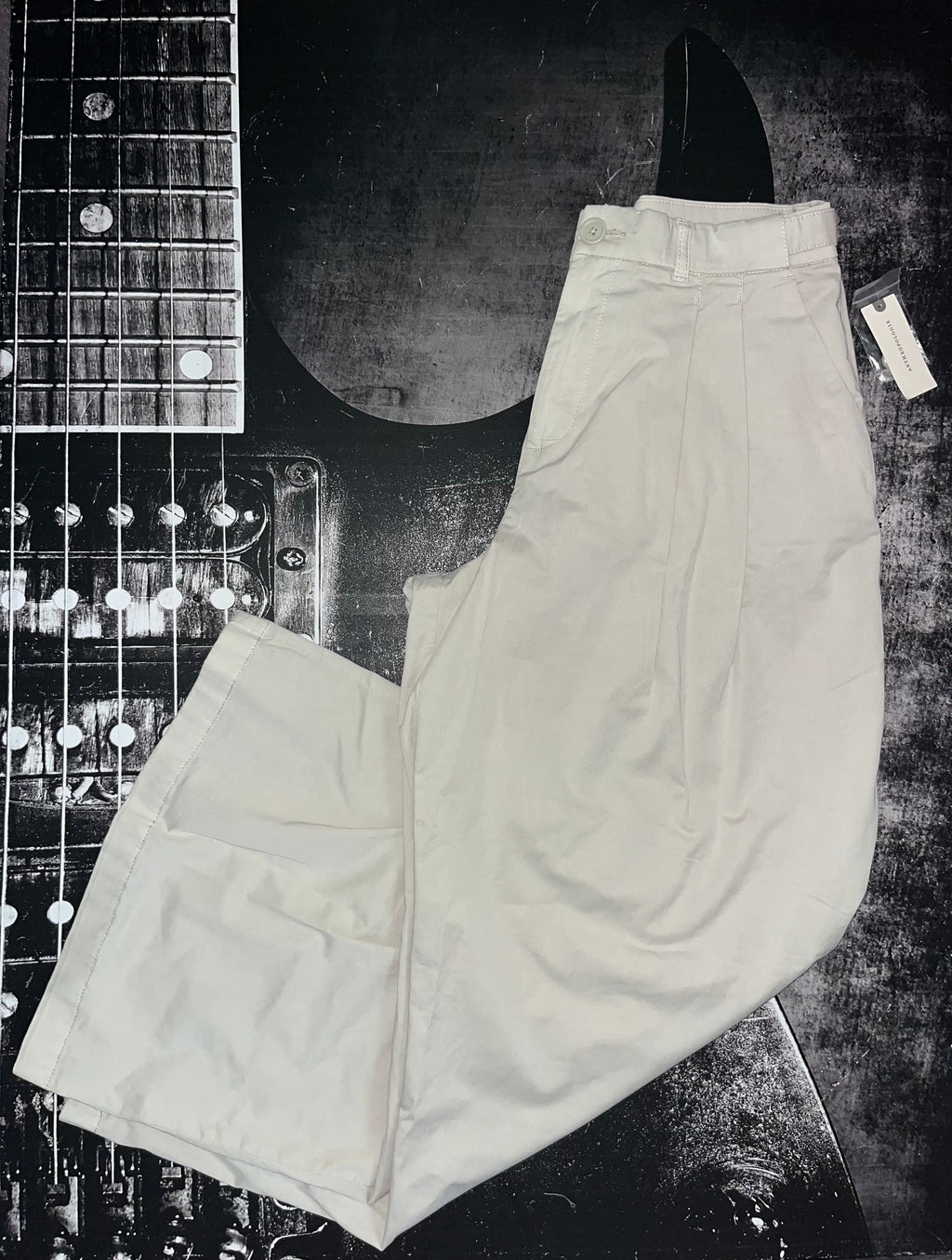 A pair of off-white or cream-colored wide-leg trousers laid flat on a dark, textured background with a guitar headstock partially visible in the corner.