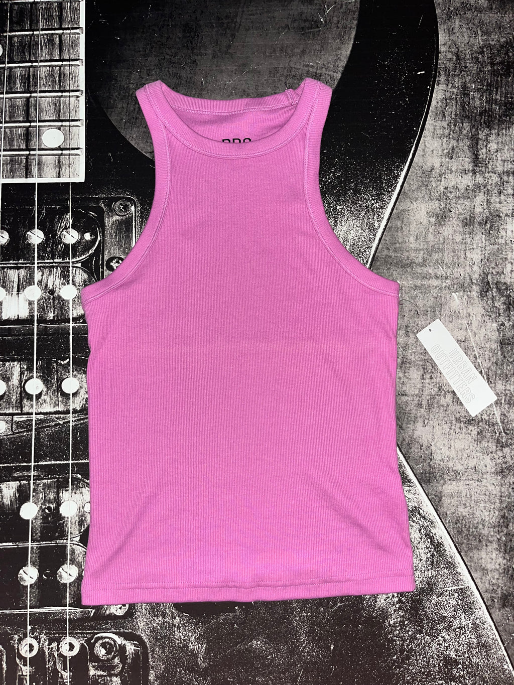 A bright pink sleeveless tank top laid flat on a black-and-white guitar against a textured gray background.