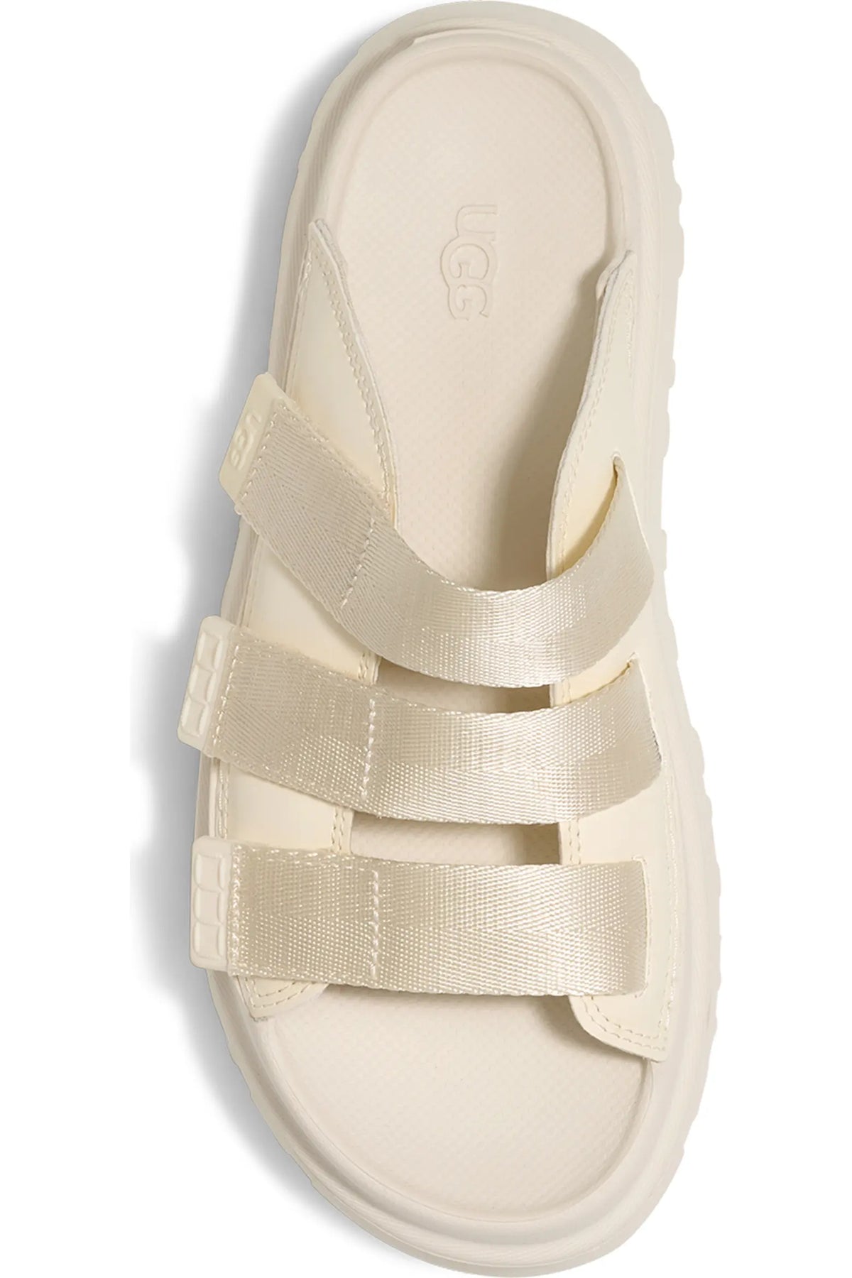 A single UGG slide sandal in a warm cream or off-white color, featuring wide elastic straps and a thick textured sole, shown from a top-down view on a white background.