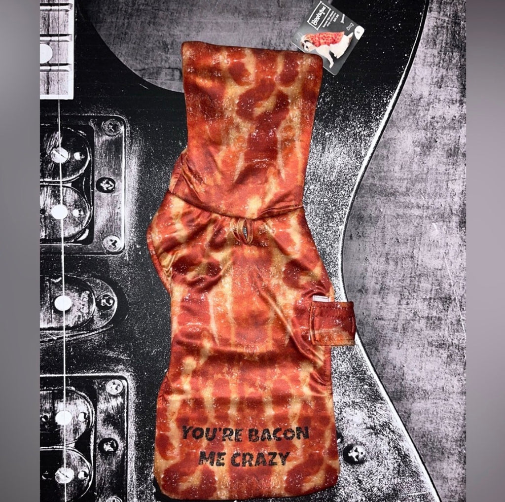 A novelty dog collar with a bacon print pattern and the text 'YOU'RE BACON ME CRAZY' printed on the front, displayed flat against a black and gray electric guitar background.