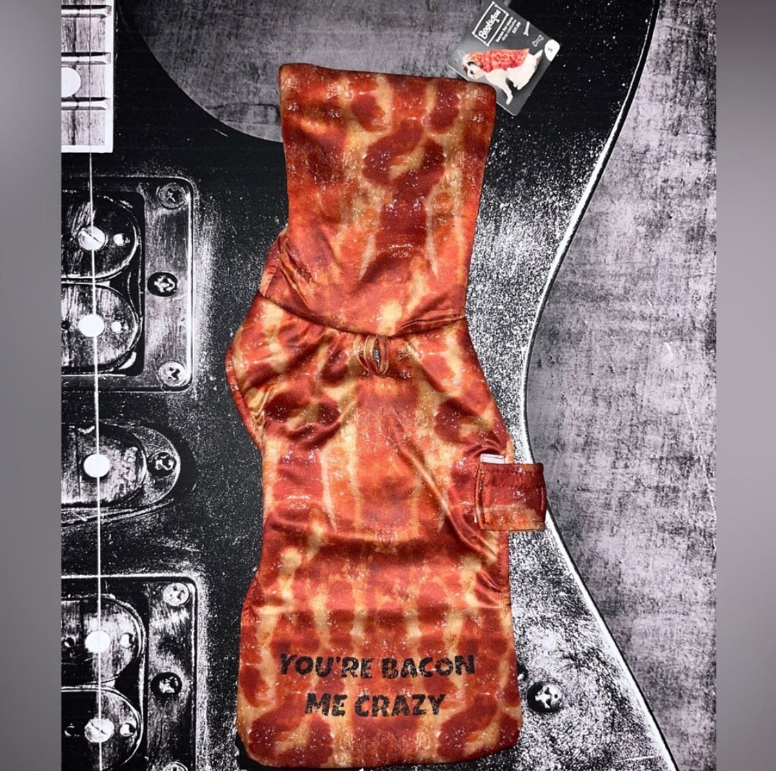 A novelty dog collar with a bacon print pattern and the text 'YOU'RE BACON ME CRAZY' printed on the front, displayed flat against a black and gray electric guitar background.