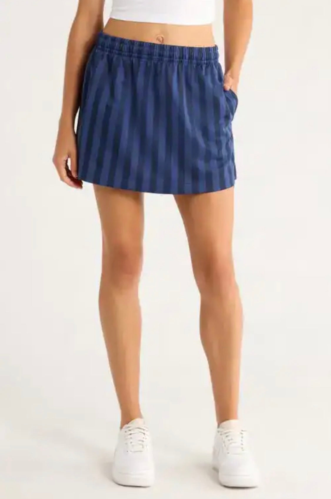 A model wearing a blue and navy vertically striped athletic mini skirt with side pockets, paired with a white crop top and white sneakers, against a plain white studio background.