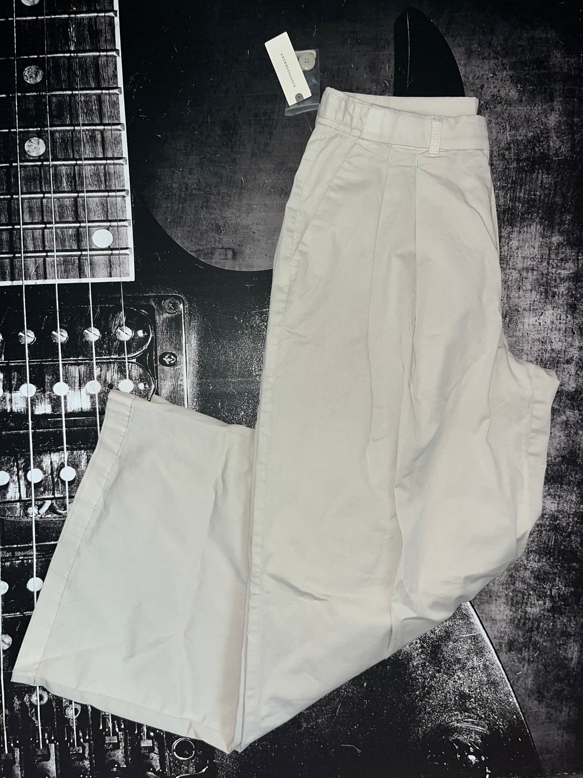 A pair of off-white or cream-colored trousers laid flat on a dark, textured surface next to a black-and-white guitar. The pants are shown with a small white retail tag attached at the waistband.