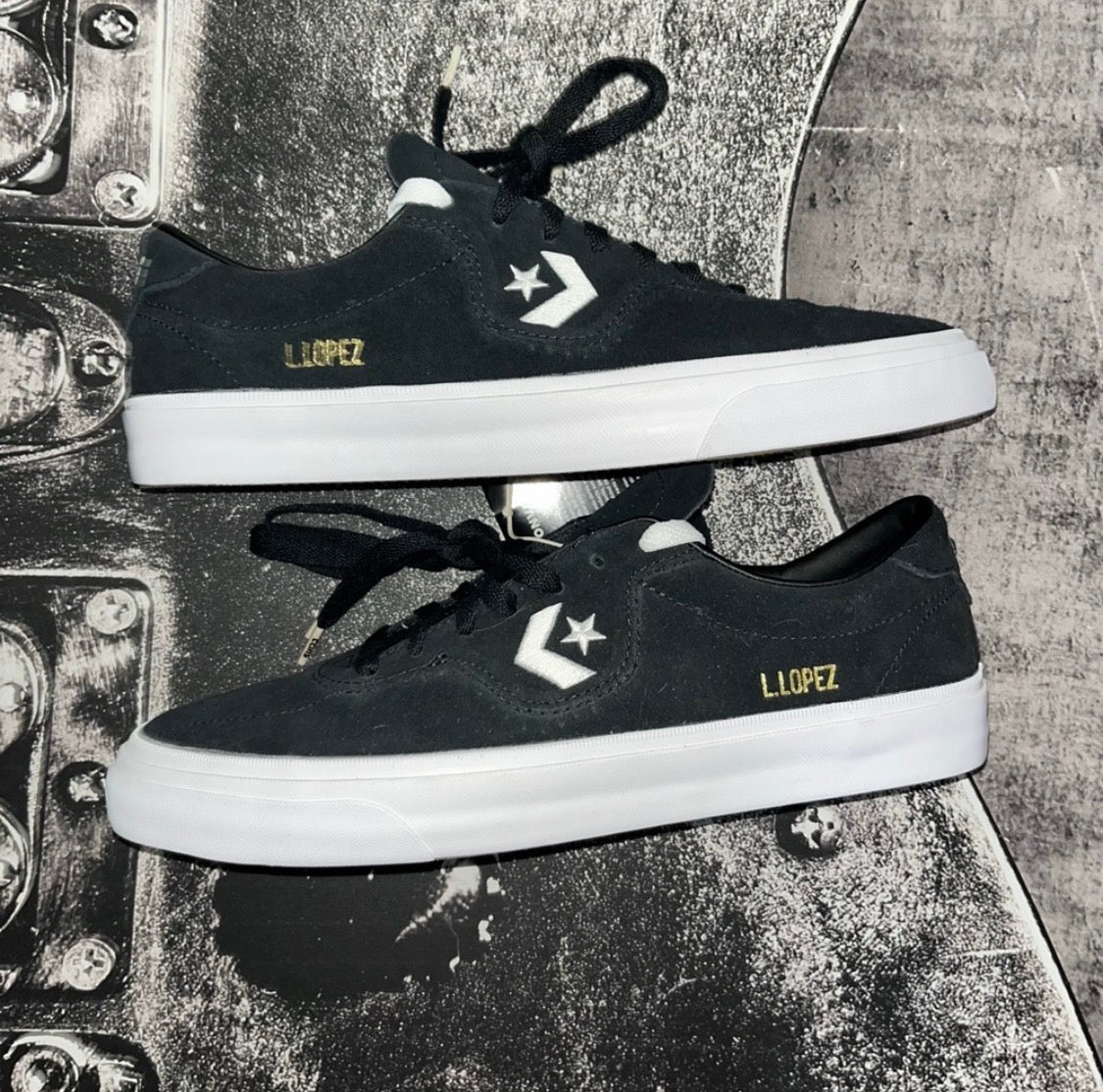 A pair of black Converse low-top sneakers with white rubber midsoles, white star logos, gold 'LOPEZ' text on the sides, and black laces, displayed on a textured metallic surface.