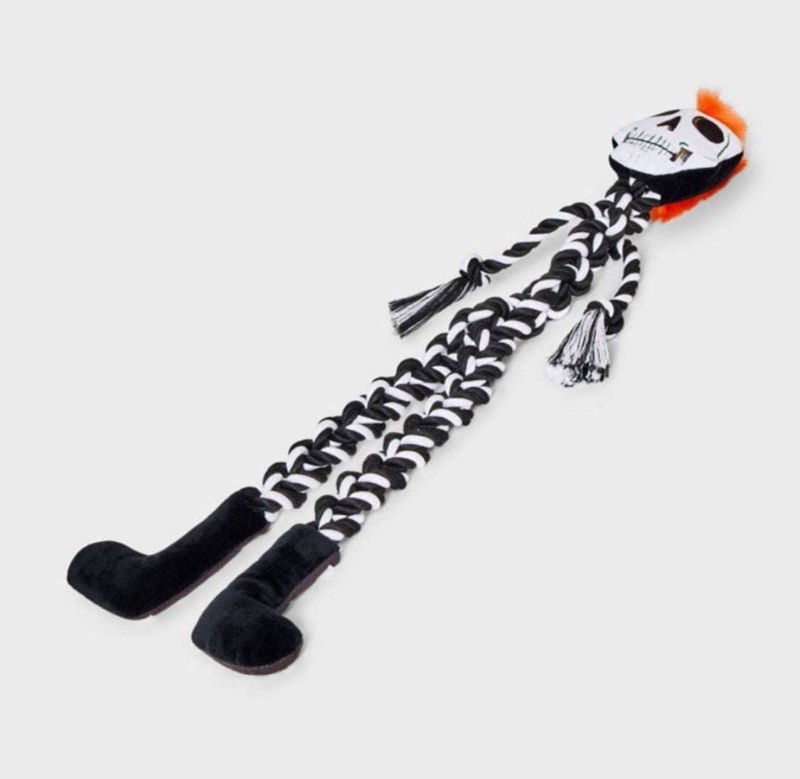 A skeleton-themed dog toy with a white skull head, black braided body, black feet, and orange hair accents, shown on a light gray background.