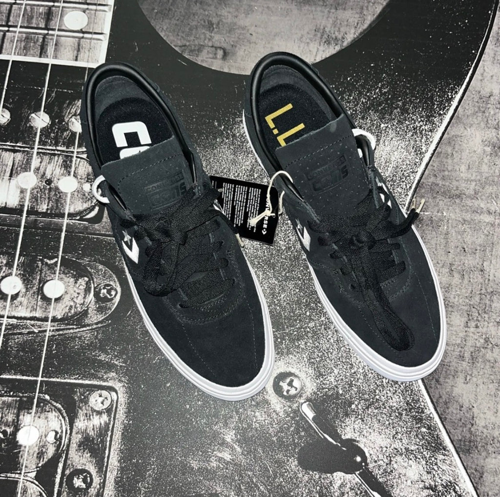 A pair of black skate shoes with white soles, laid flat on a guitar headstock. The shoes feature a hook-and-loop strap closure with a white logo, black laces, and a black hangtag attached.