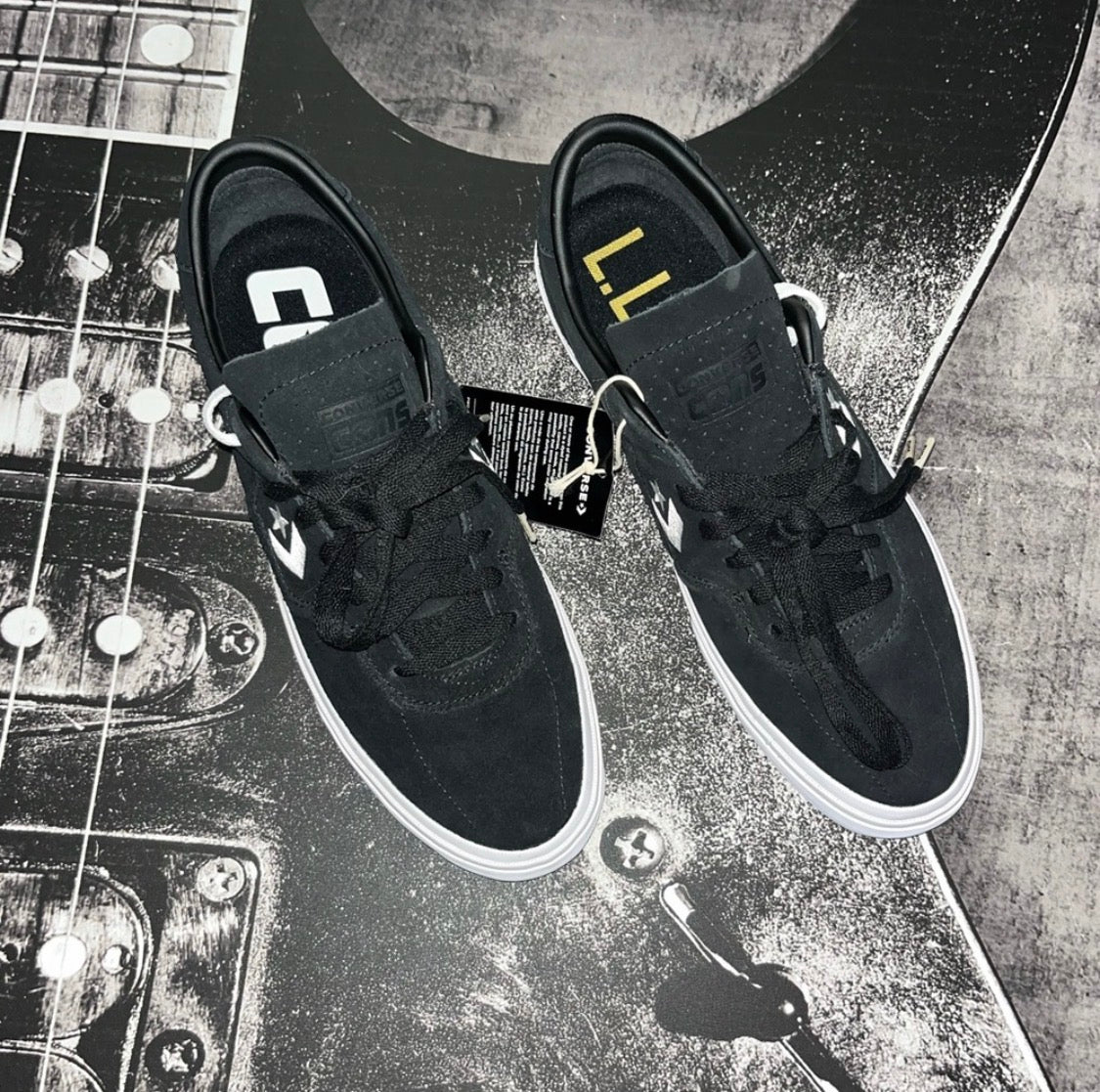 A pair of black skate shoes with white soles, laid flat on a guitar headstock. The shoes feature a hook-and-loop strap closure with a white logo, black laces, and a black hangtag attached.