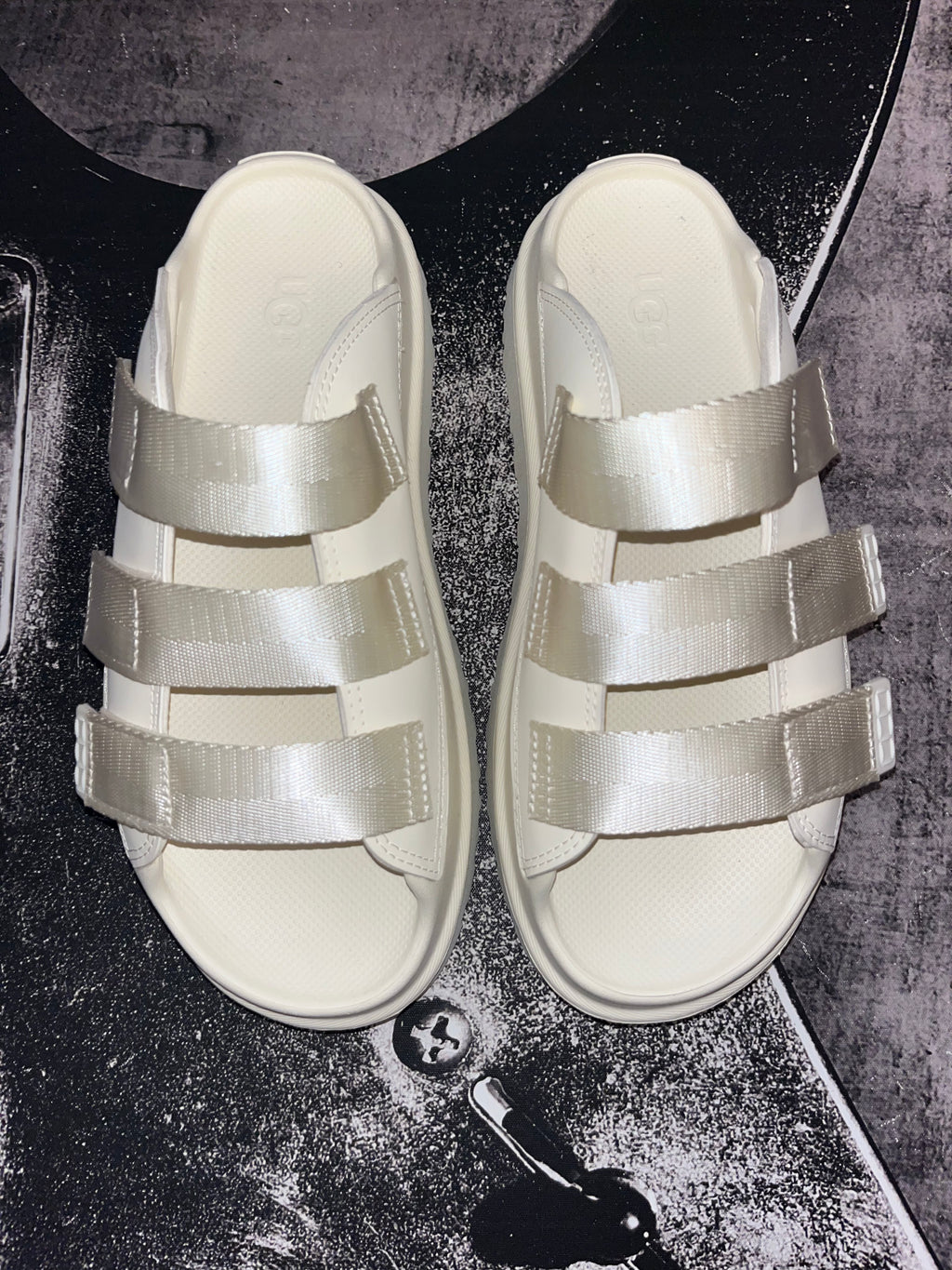 A pair of cream-colored slide sandals with wide, shiny satin-like straps, laid flat on a dark textured surface.