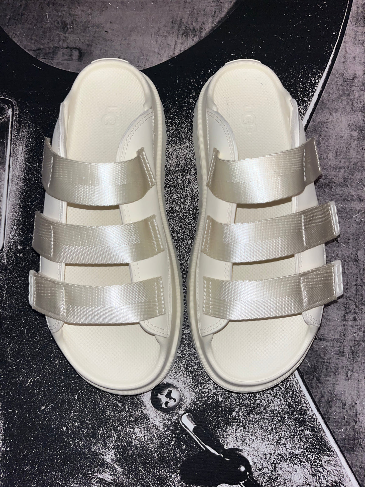 A pair of cream-colored slide sandals with wide, shiny satin-like straps, laid flat on a dark textured surface.