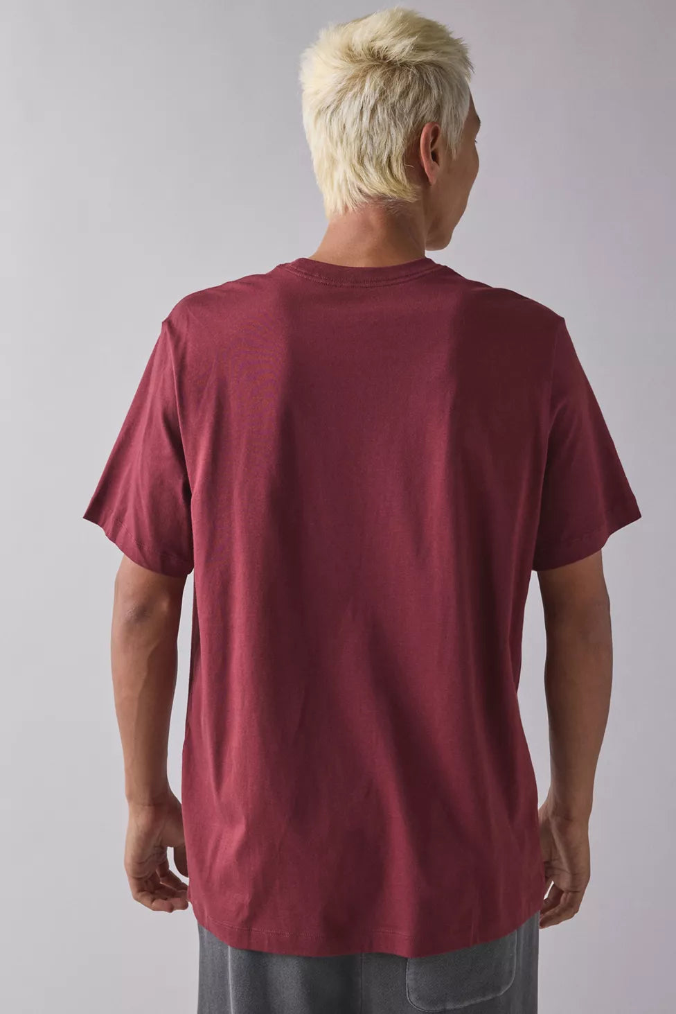 A man with short blonde hair is shown from behind wearing an oversized, dark red crewneck t-shirt and gray pants against a plain light gray background.