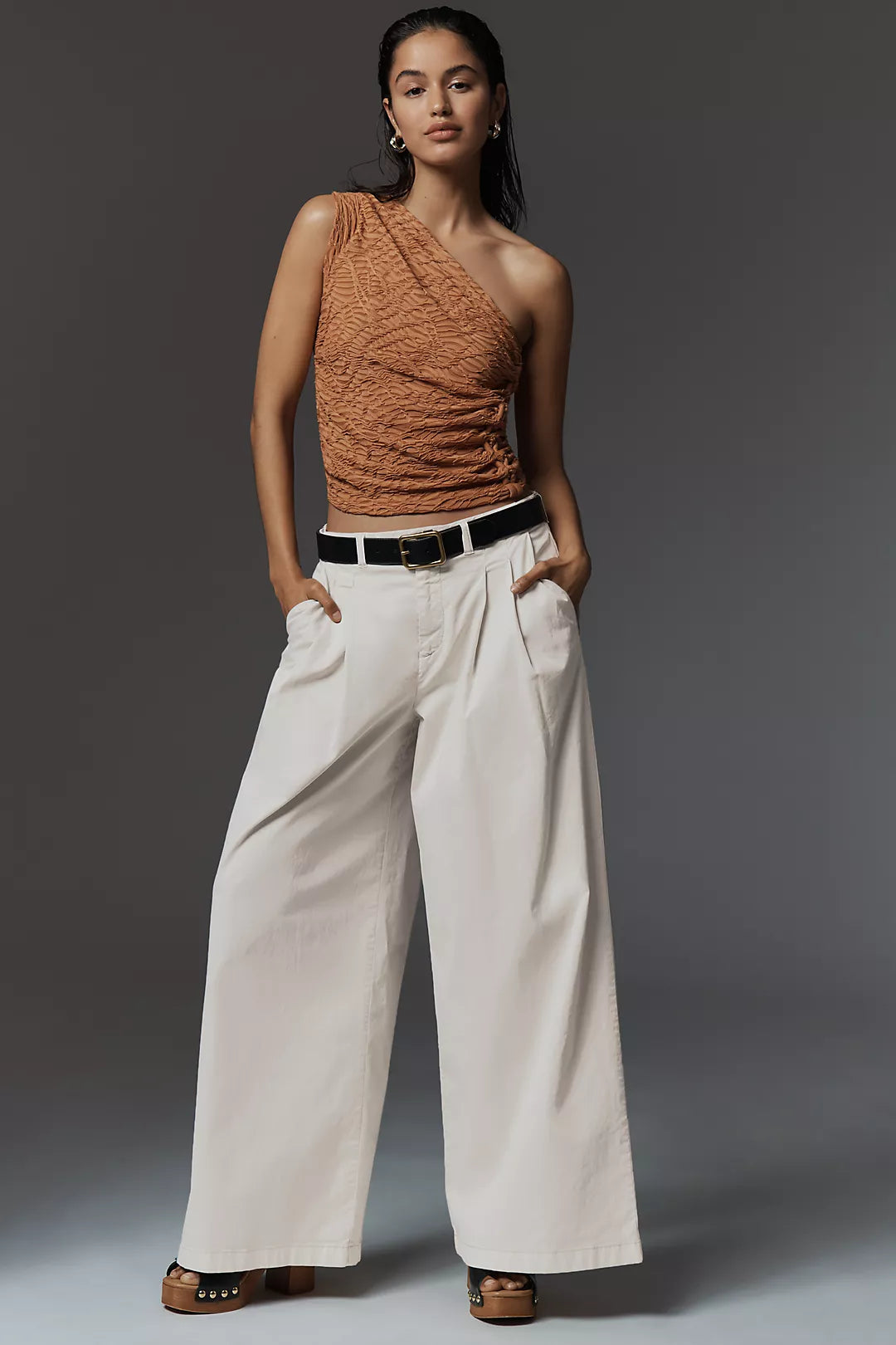 A model stands against a dark gray studio background wearing a textured rust-orange one-shoulder top, off-white wide-leg trousers with a black belt, and brown sandals.