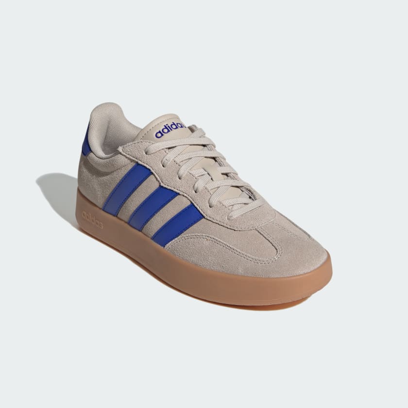 An Adidas low-top sneaker in beige suede with blue three-stripes, a beige tongue, and a gum sole, shown on a light gray background.