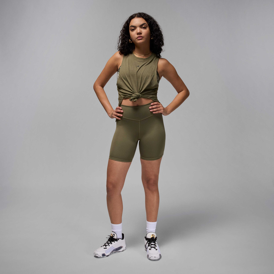 A model stands against a light gray studio background wearing an olive green athletic set consisting of a sleeveless cropped top with a front knot detail and matching high-waisted biker shorts. She is also wearing white socks and white sneakers with black details.
